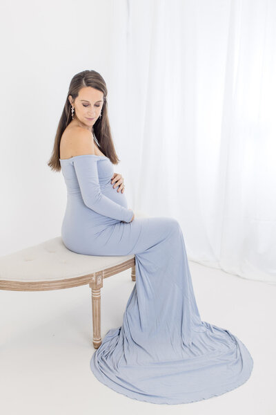 A pregnant woman sitting on a bench in a white studio wearing a blue dress