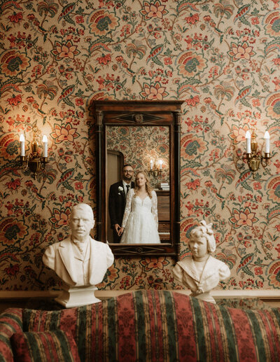 A creative shot of a bride and groom reflected in a vintage mirror surrounded by ornate wallpaper and antique busts.