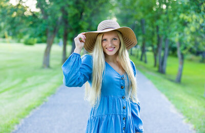 High School senior girl wearing blue dress and tan hat standing on a bike path in the middle of summer.