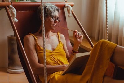 Middle aged, light skinned woman in a sleeveless yellow romper sits reading in a brown leather hammock chair. Photo by Los Muertos Crew via Pexels.