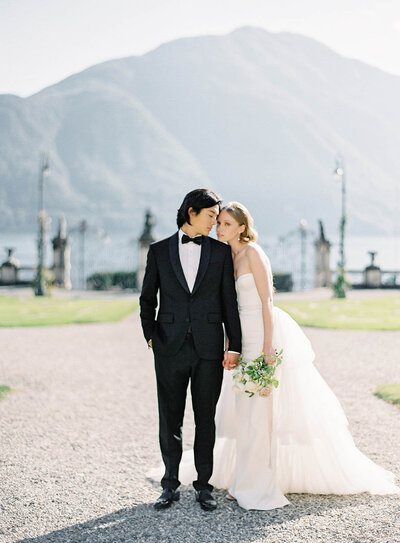 Bride and groom standing facing each other with his arms around her waist and her arm around his neck and the other down to her side holding a bouquet. They are in front of the ceremony site that is in tall grasses with a horse shoe shape of layers of flowers from ground height up to 4 feet tall.  Massive delilahs throughout the florals. Behind them are mountains of evergreens then bare mountains beyond that. Blue skies with some clouds.  She is wearing a uniquely designed colorful wedding gown with clay embellishments. He is in a navy blue tuxedo. In the Colorado Rockies. Photographed in Estes Park by wedding photographers in Charleston Amy Mulder Photography