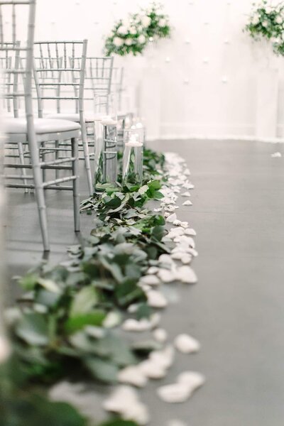 A close-up of the ceremony aisle at SoHo63, a Chandler wedding and event venue, lined with white rose petals, lush greenery, and glass candle holders beside silver chiavari chairs—creating a romantic, modern atmosphere.