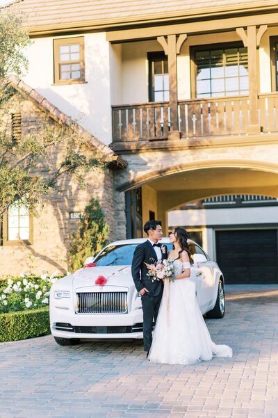 Kohnur elegant bride with soft glam makeup and long loose waves posing with groom and a luxury Rolls Royce at a modern estate.