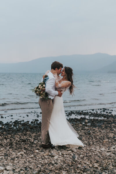 groom holding bride around the waist while bride holds groom around the neck with faces almost touching standing on a rocky beach in Lake Tahoe by California elopement photographer Kasey Mantiply