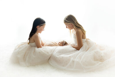 2 sisters in pale pink dresses holding their newborn baby sister wrapped in a peach swaddle taken on white background.