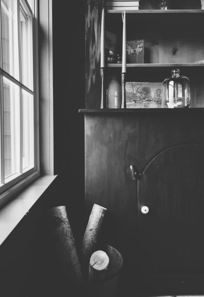 black cabinet with candlesticks, glass cloche antique art sketch and a tin bucket with birch logs in front of the cabinet