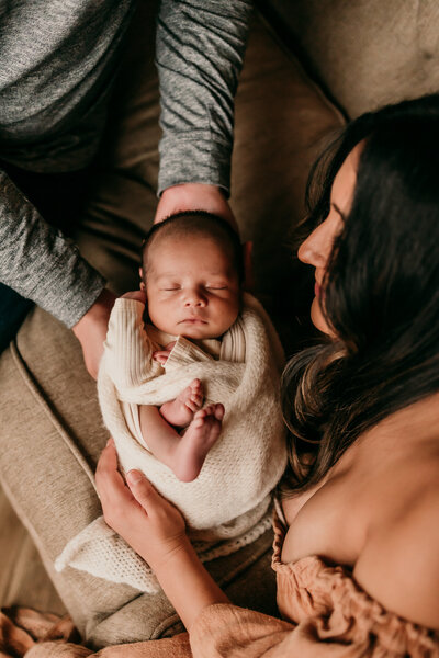 Newborn baby wrapped in a cream swaddle resting in the arms of both parents during a cozy in-studio lifestyle session, with mom wearing an off-shoulder rust-colored dress

