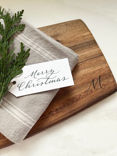 Wood cutting board with wood-burned monogram letter M and Merry Christmas gift tag in dark green calligraphy