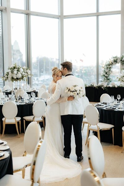A bride and groom stand in the middle of the O'Born Room at the National Arts Centre for their wedding, featuring a beautiful white dress and white and black tuxedo, surrounded by black guest tables and luxurious white and gold leather chairs with white floral centrepieces in the glass room.
