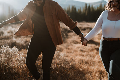 Couple holding hands and walking through a sunlit field during their playful Breckenridge engagement session.