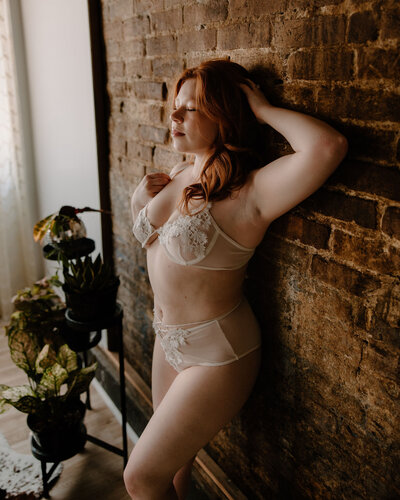 Dark moody aesthetic boudoir photography with woman leaning against brick wall in white 2 piece lingerie with her hand on her hair, plants and hardwood floor in the background captured by Morgan Ashley Lynn Photography, photographer for Lake Country, Waukesha and Milwaukee, WI