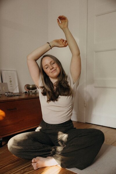 Eine Frau ist im Yogasitz Lotus und räkelt lächelnd die Arme in die Luft