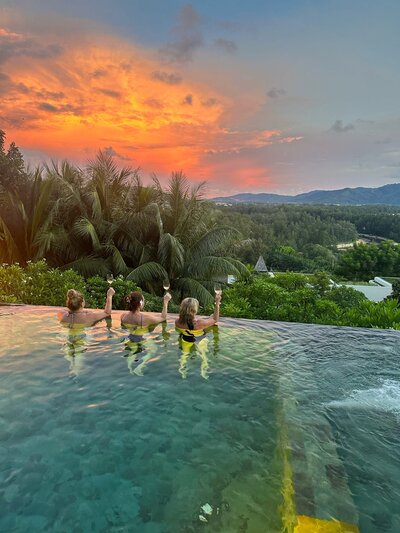 Three women relax in an infinity pool at sunset, raising their arms in celebration. The pool overlooks lush greenery and a distant mountain range, with the sky glowing in vibrant orange and pink hues.