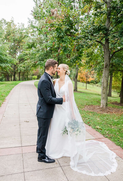 A bride stands next to a groom in a park in cincinnati ohio. It's fall and the trees are turning yellow. They are smiling at each other with noses touching.