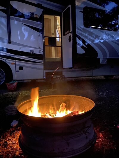 A campfire burns brightly in a metal fire pit at night, set before a lit-up RV with the door open. Shadows cast intriguing patterns on its exterior, creating a serene camping scene at one of the inviting Mayo Florida campgrounds.