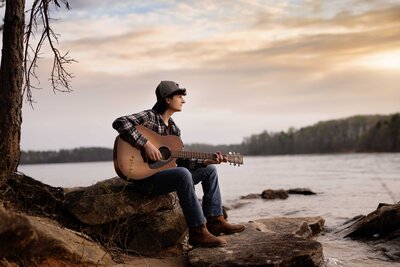 Senior photoshoot by the lake with boy grad sitting on rock and playing guitar, looking into the sunset. Graduation photo taken by Jenni Nikki photography in Atlanta, GA.