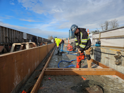 An operator on a site operating heavy machinery to drill holes into existing concrete and inserting steel dowels to transfer loads and prevent cracking at joints.