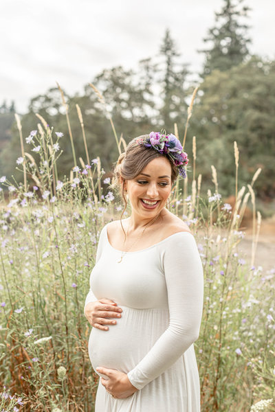 Pregnant woman in white off the shoulder dress wearing a floral crown and smiling as she is looking down over her shoulder stands in a field of wildflowers during her outdoor maternity photography session in Oregon.