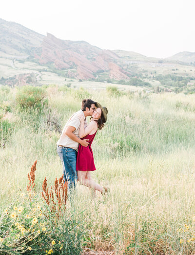 denver couples photography with man holding womans waist and leaning in to kiss her and she giggles and smiling while wearing a red dress in the mountains