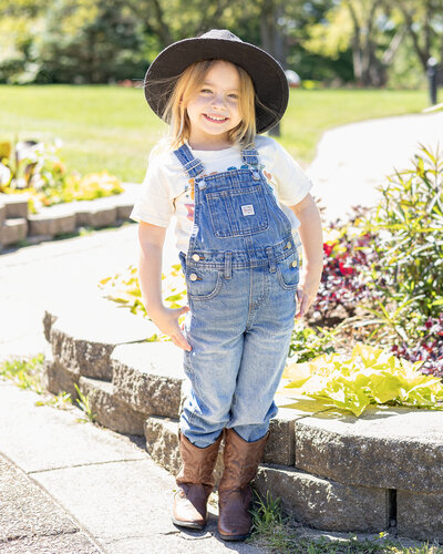 Young blonde girl wearing blue denim overalls, black hat, at the park, smiling at the camera