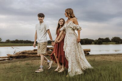 A woman and two children walk hand-in-hand on grass near a lake, with logs and trees in the background under a cloudy sky.