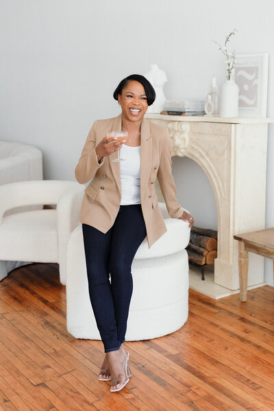 Dark skinned woman with short hair leans on white chair and laughs while holding  a glass of champagne. Background is fireplace and white walls