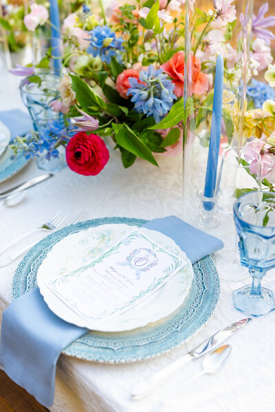 A close-up of an elegant spring table setting with a blue and white color palette, featuring a floral centerpiece, blue glassware, blue taper candles, and a decorative menu placed on a layered charger and plate with a blue napkin.