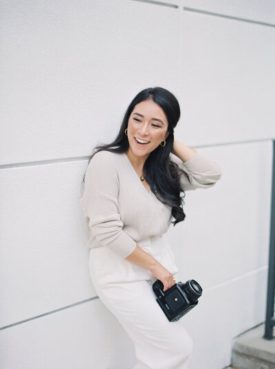 DFW wedding photographer, Stephanie, leaning against a white wall holding her camera wearing white dress pants and a cream sweater
