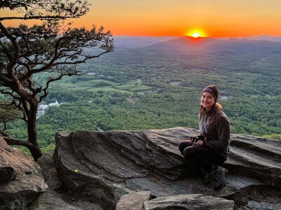  photographer sits on a rock on top of a mountain holding her camera and smiling. The sun is setting behind a mountain range turning the sky a bright orange.