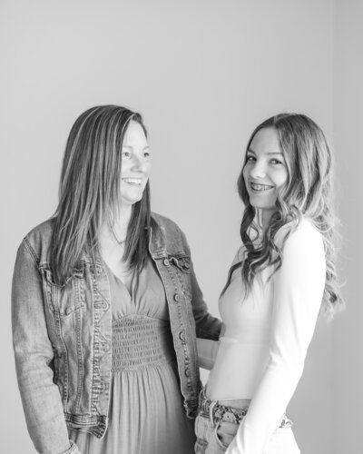 black and white photo of a mother and daughter in a studio, mom looking at daughter smiling while daughter is looking at the camera.  Each have long hair