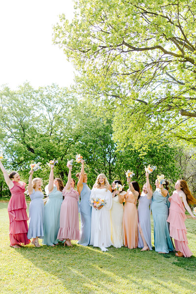 Bride surrounded by bridesmaids in mismatched pastel gowns, holding bouquets and celebrating under a sunlit canopy of trees during a spring wedding in Oklahoma.
