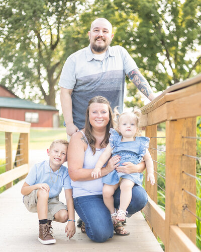 Family of four on a bridge in a wildlife sanctuary.  Dad is standing up behind his family wearing a striped t-shirt, has a shaved head with a dark brown beard, smiling at the camera.  Son and mom are knelt down with toddler daughter on her lap.  All looking at the camera smiling.  Son has short light brown hair, light blue collared short sleeve shirt, and khaki shorts.  Mom has long brown hair wearing blue jeans and white spaghetti strapped shirt.  Daughter has blonde pigtails and wearing a blue and white dress.