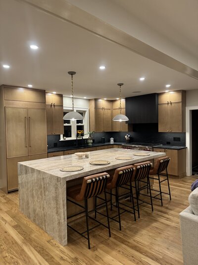 Modern kitchen renovation with waterfall stone island, wood cabinetry, black range hood, and leather bar stools.