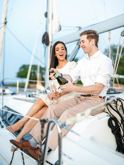 A couple sits on a sailboat, smiling and celebrating. The man is pouring champagne into glasses while the woman holds hers, looking excited. Both are dressed in light, summery clothes, with the backdrop of calm water and a clear sky.