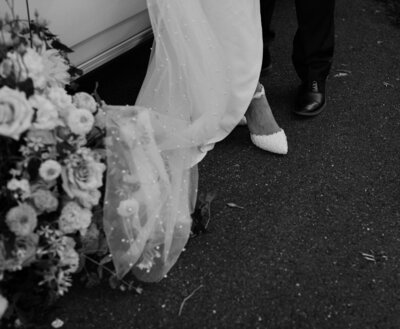 A black and white photo of a white lace beaded bridal veil draped on the ground and over a creation of flowers.