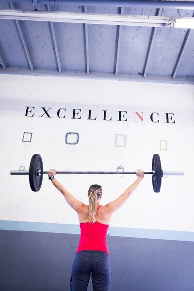 A female fitness trainer holding a barbell over her head, wearing a red tank top, and the word excellence on the wall behind her.