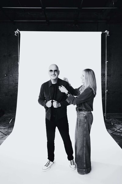 black and white image of a man smiling in sunglasses while a woman adjusts his jacket during a styling session
