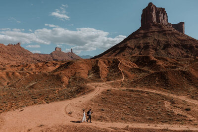 bride and groom walking down a dirt road in the desert with mountains and large rock formations in the background 