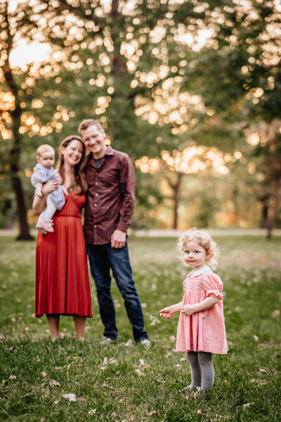 A young family of four standing in a park at sunset. The parents smile in the background, holding a baby, while a toddler in a pink dress holds flowers in the foreground, looking at the camera.