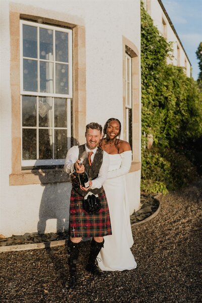 An LGBTQ+ couple candidly pose together for wedding photography in a garden. They both wear dresses and smile.