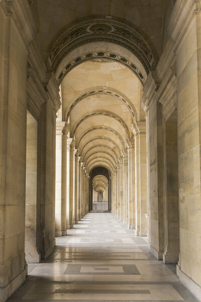 A long, arched corridor with ornate stone arches and columns lining both sides. The floor has a geometric pattern, and the perspective creates a tunnel-like view, receding into the distance. Soft lighting highlights the architectural details.