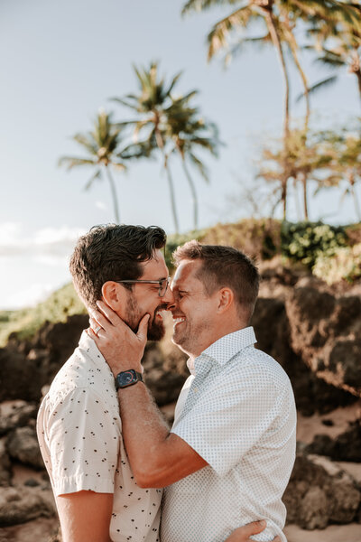 two gentleman about to kiss during their engagement shoot on a beautiful beach in Maui with rocks, sand and palm trees in the background.