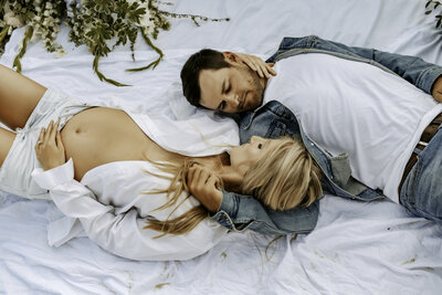 Family Photographer, a couple, man and woman lay on white sheets together and gaze into each other's eyes
