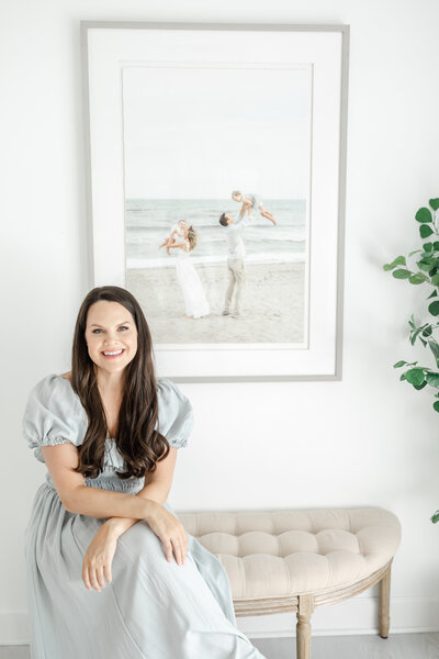 Connecticut family photographer, Kristin Wood, sits on an upholstered bench beneath a framed family portrait