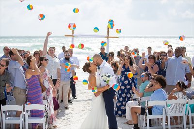 Couple shares a kiss at the end of the aisle as guest throw mini beach balls and cheer at the end of a beach wedding.