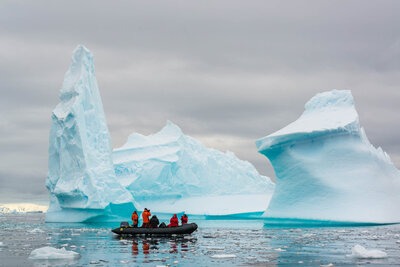 A small boat with people in bright jackets floating near large icebergs in icy waters under a cloudy sky.