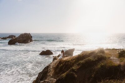 A couple embraces on a rocky cliffside overlooking the Pacific Ocean during their Big Sur elopement, with waves crashing below and golden sunlight reflecting off the water