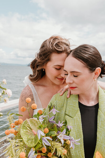 Bold, colorful wedding bouquet sits on display next to bright orange strappy heels. There is backlighting coming in from the window. The bridal bouquet flowers are green, orange, and light purple. There is a chartreuse green ribbon cascading from the bouquet..