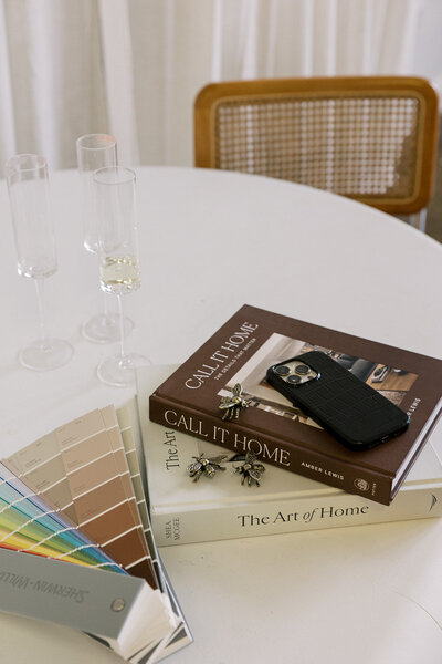 A round white table with three stacked books titled "The Art of Home," "Call It Home," and a third book. A smartphone, bee-shaped accessories, two empty champagne glasses, and a color palette fan are on the table. A cane chair is in the background.