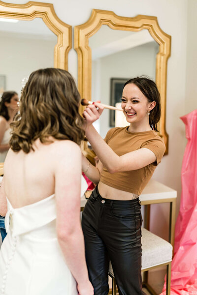 A smiling makeup artist wearing a brown crop top and black pants applies makeup to a woman in a white strapless dress in front of two gold-framed mirrors.